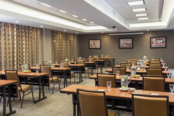 A hotel conference room with multiple tables and chairs, equipped with water pitchers, glasses, and small plates of food.