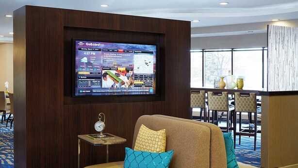 A hotel lobby with a large flat-screen TV displaying news, a comfortable seating area with a sofa and cushions, a small table with a clock, and a view of the dining area through large windows.