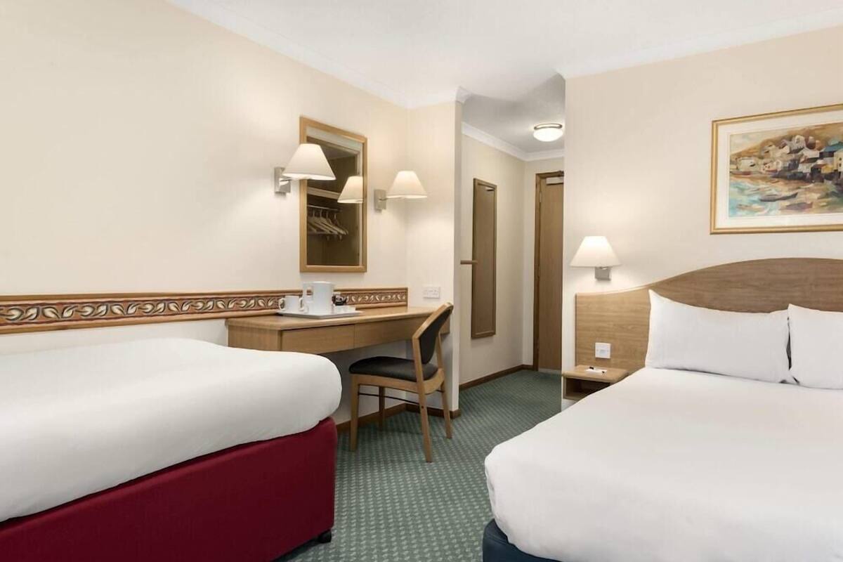 A hotel room with two beds, a desk with a chair, a mirror, a painting on the wall, and a door leading to another room.