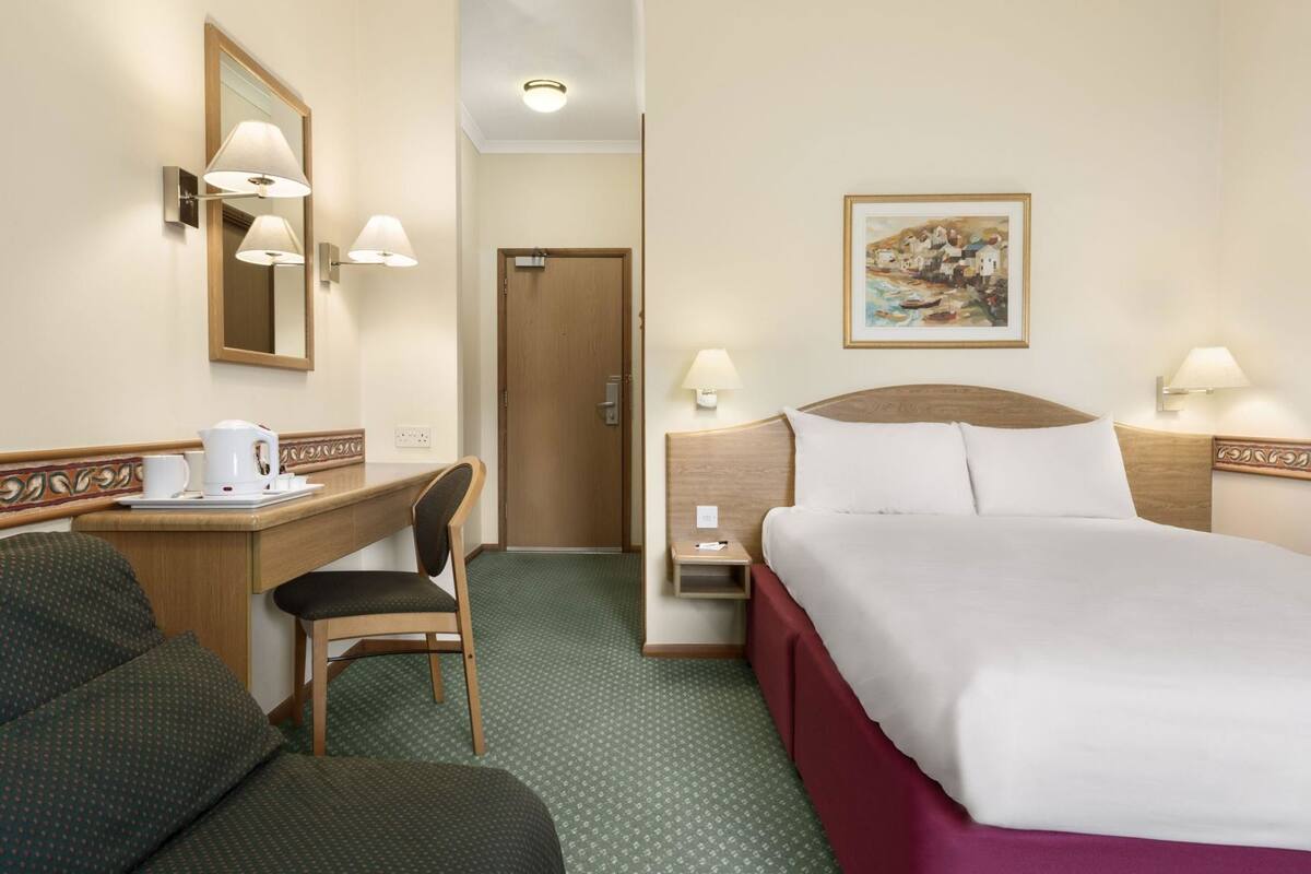 A hotel room with a bed, a desk with a chair, a mirror, and a painting on the wall.