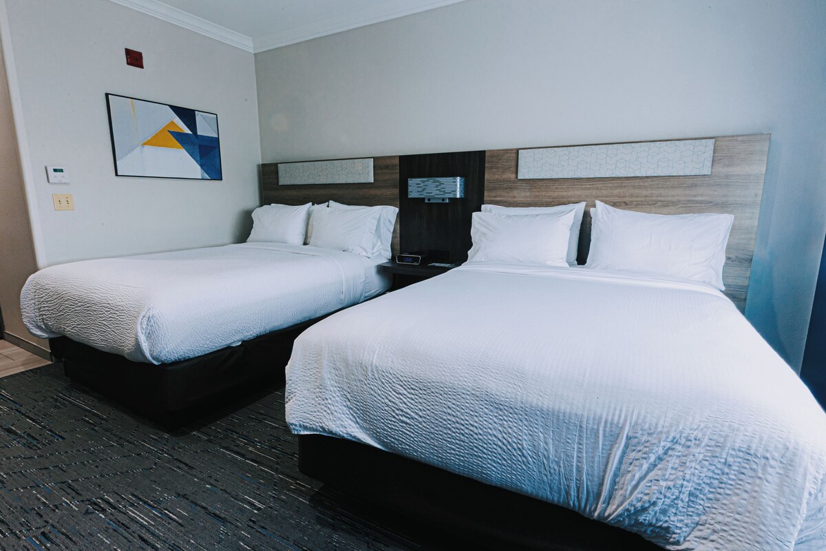 Quarto de hotel com duas camas, uma mesa de cabeceira, um relógio digital e um termostato fixado na parede.