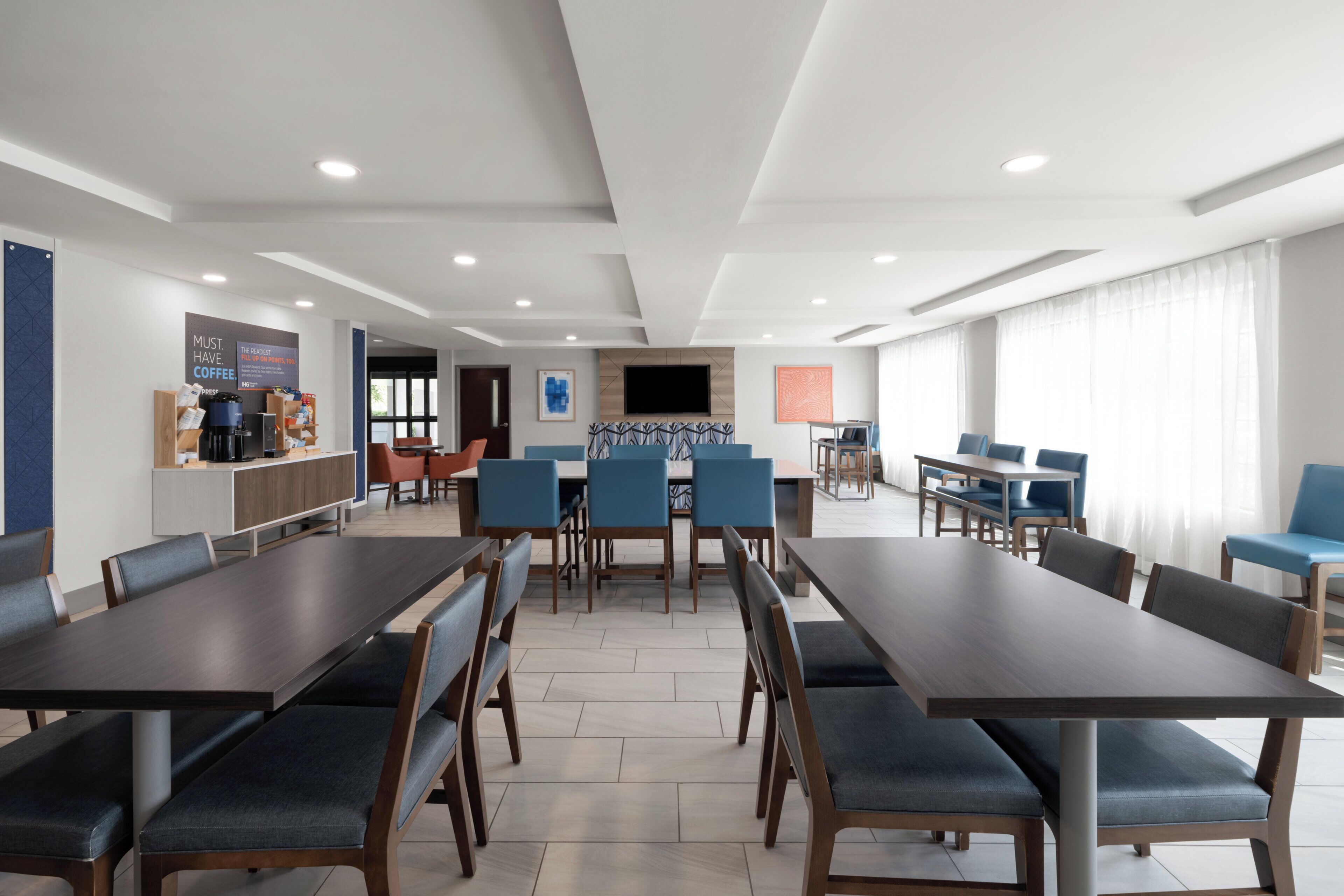 A hotel dining area with multiple tables and chairs, a coffee station, and a TV mounted on the wall.