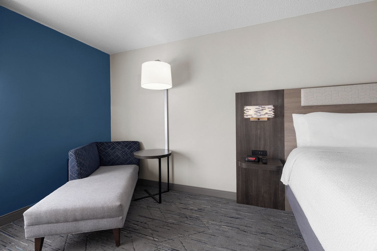 A hotel room with a bed, a small sofa, a nightstand, a lamp, and a wall-mounted clock.