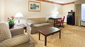 A hotel room with a patterned sofa, a small table, a red chair, a desk with a lamp, a microwave, and a painting on the wall. - Holiday Inn Exp Winston Salem by IHG (Winston-Salem)