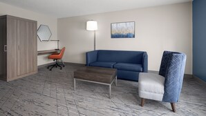 A hotel room with a wooden wardrobe, a blue sofa, a grey armchair, a wooden coffee table, a floor lamp, and a painting on the wall. - Holiday Inn Exp Winston Salem by IHG (Winston-Salem)