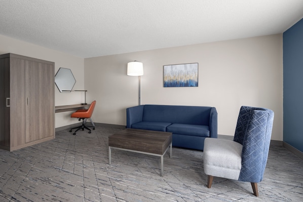 A hotel room with a wooden wardrobe, a blue sofa, a grey armchair, a wooden coffee table, a floor lamp, and a painting on the wall. - Holiday Inn Exp Winston Salem by IHG (Winston-Salem)
