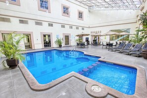 An indoor swimming pool with lounge chairs and umbrellas, surrounded by potted plants and a tiled floor.
