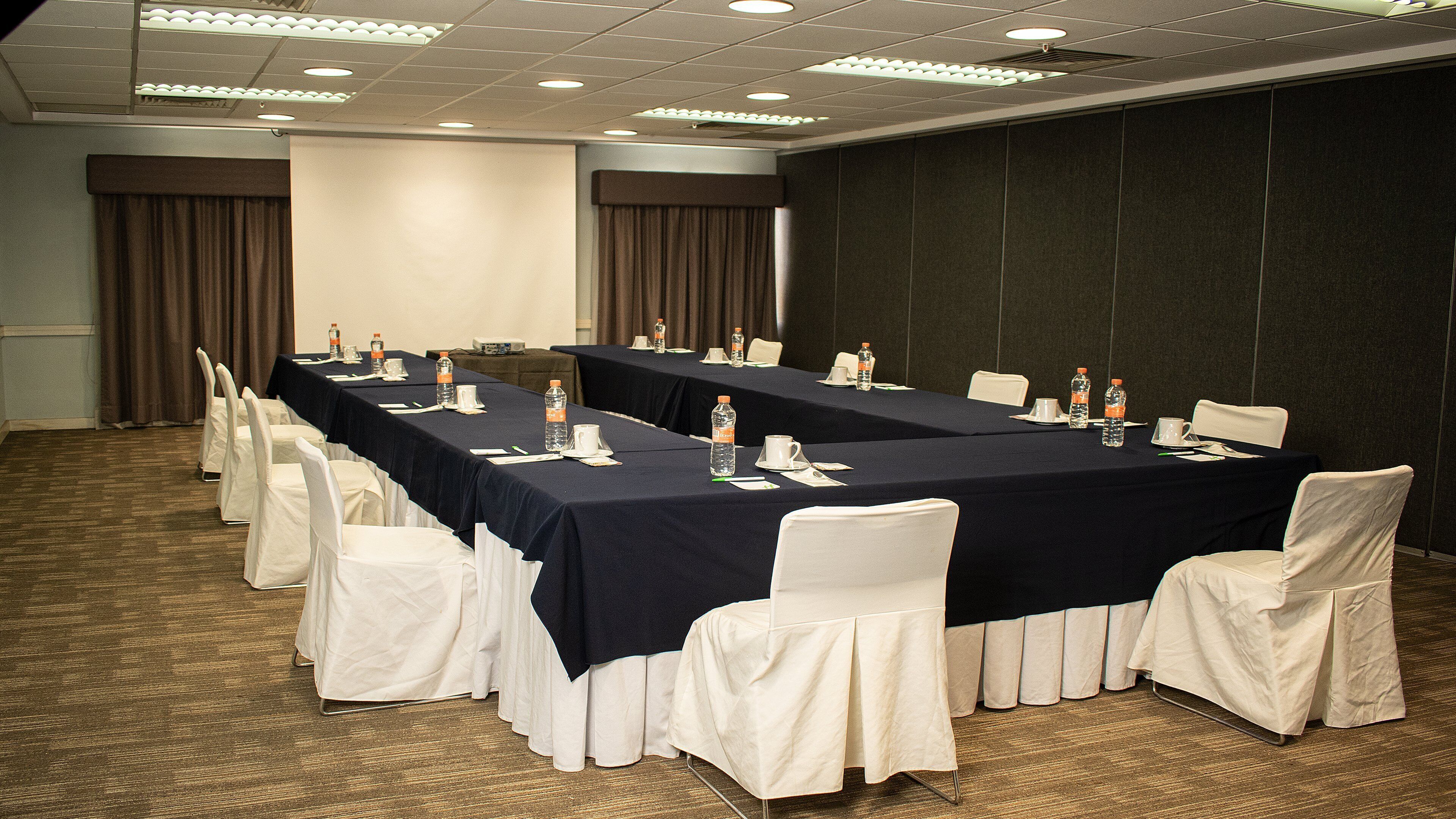 Un salón de conferencias con una larga mesa, sillas blancas y botellas de agua.