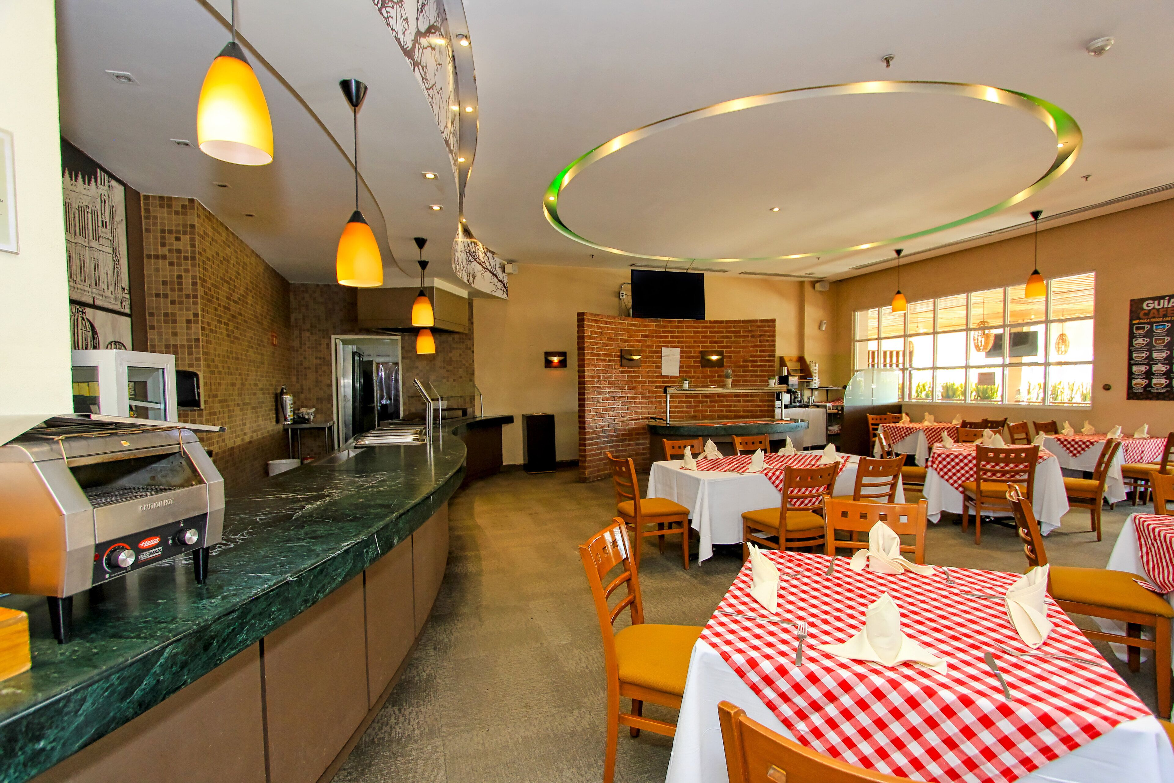 A restaurant with a kitchen area, tables set with red and white checkered tablecloths, and pendant lighting.