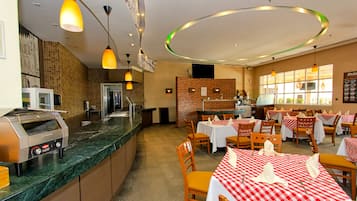A restaurant with a kitchen area, tables set with red and white checkered tablecloths, and pendant lighting.