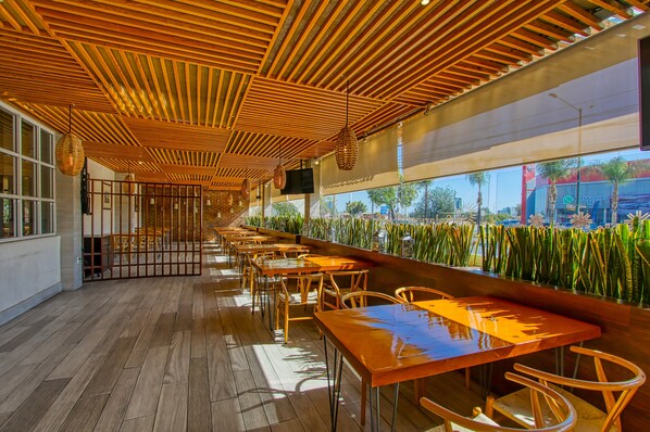 A long wooden bar with multiple tables and chairs, a wooden ceiling with slats, and large windows with a view of palm trees and buildings outside. -  Holiday Inn Leon - Poliforum by IHG (León)
