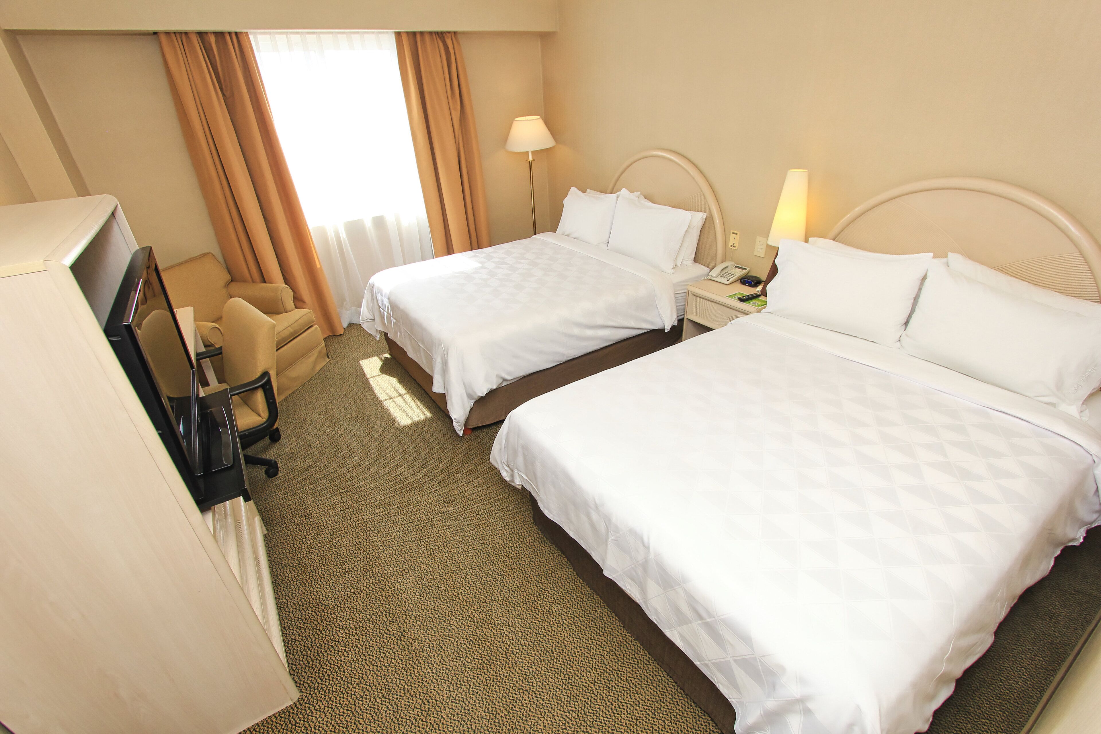 A hotel room with two beds, a desk, a chair, and a lamp.