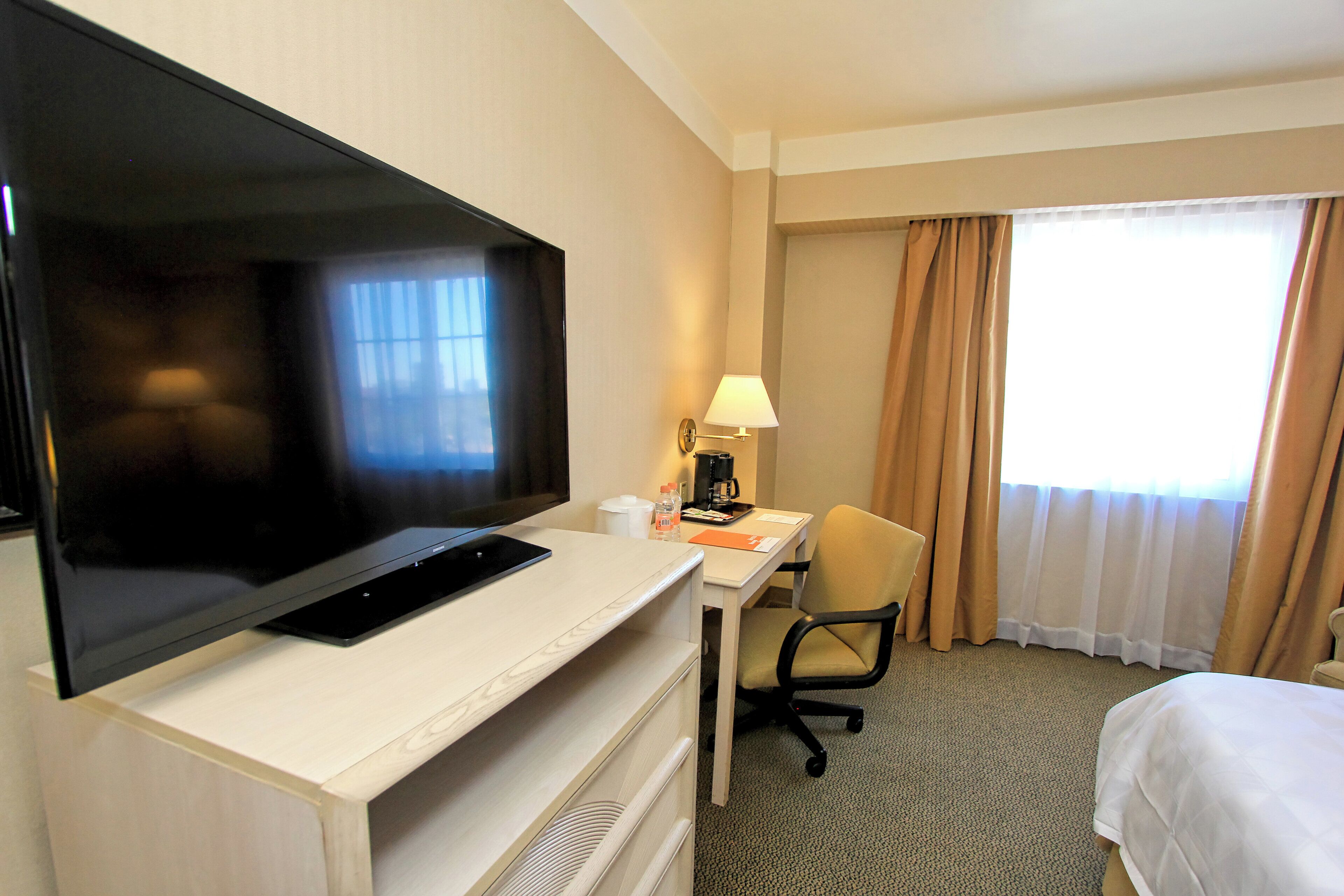 A hotel room with a flat-screen TV on a desk, a chair, a bed, and a window with curtains.