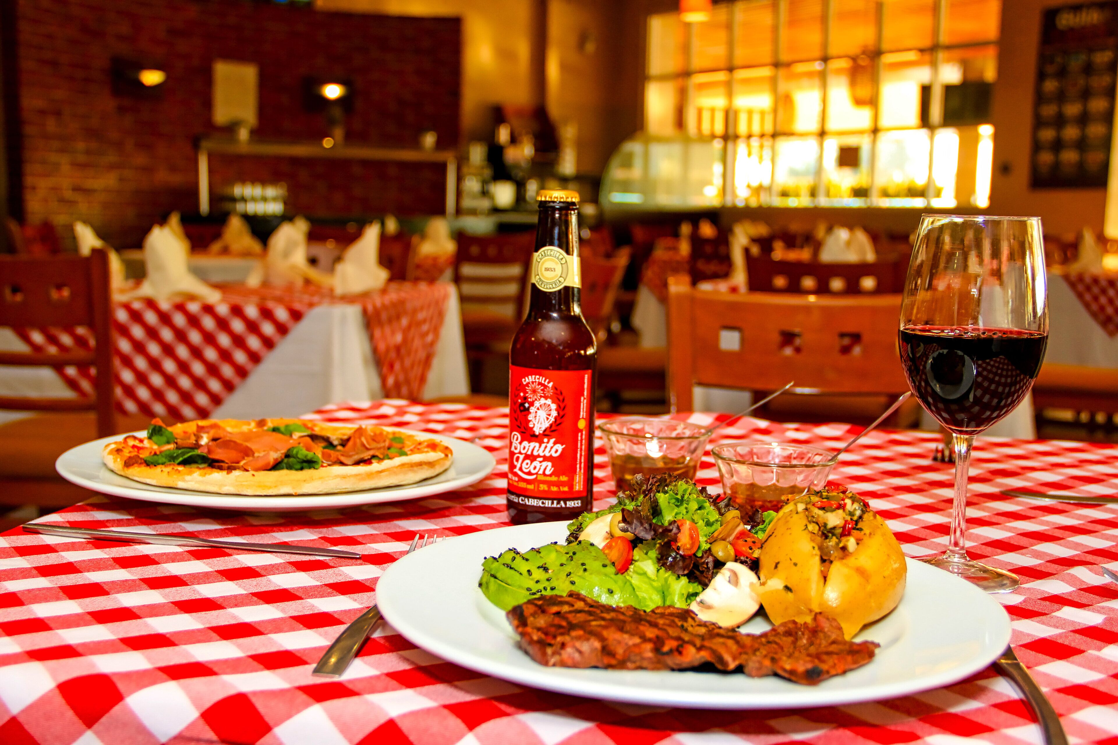 Une table de restaurant recouverte d’une nappe à carreaux, une bouteille de bière Bonito León, un verre de vin rouge et des assiettes remplies de nourriture, notamment une pizza et une salade.