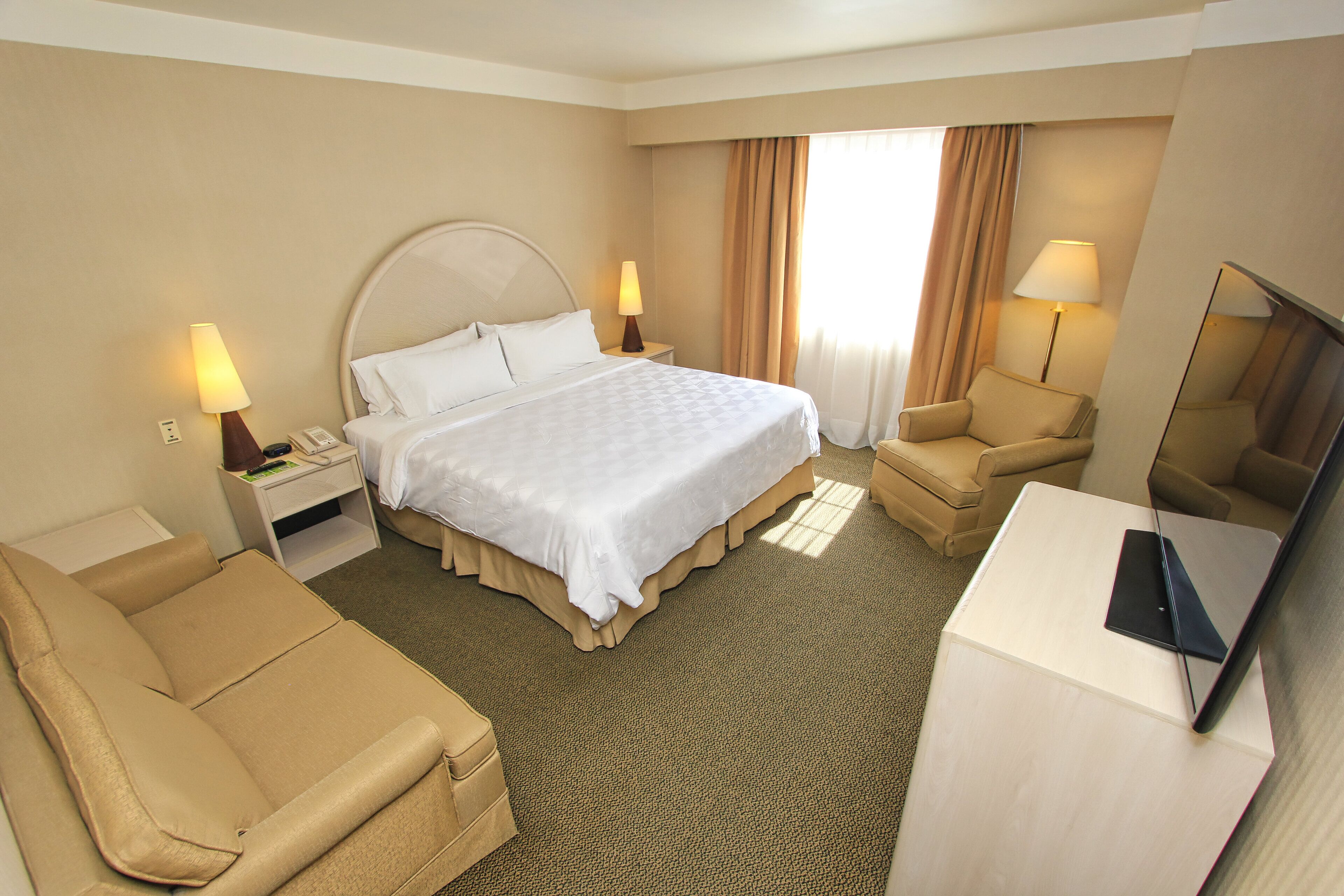 A hotel room with a bed, two bedside tables with lamps, a sofa, a desk with a laptop, and a TV.