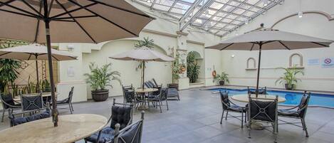 Un área de piscina interior con varias mesas y sillas, rodeada de plantas en macetas y sombrillas.
