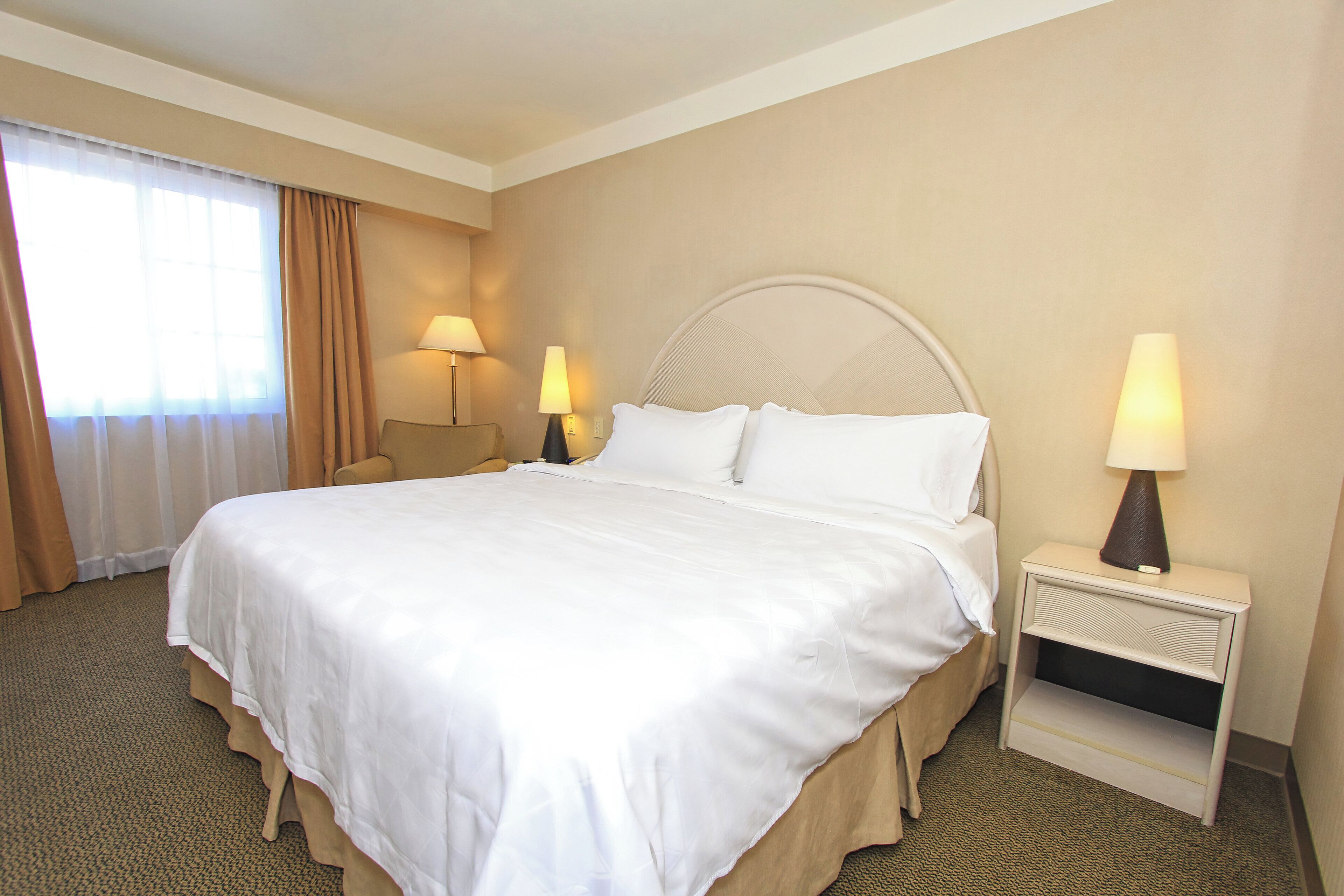 A hotel room with a large bed, two bedside tables with lamps, a chair, and a window with curtains.
