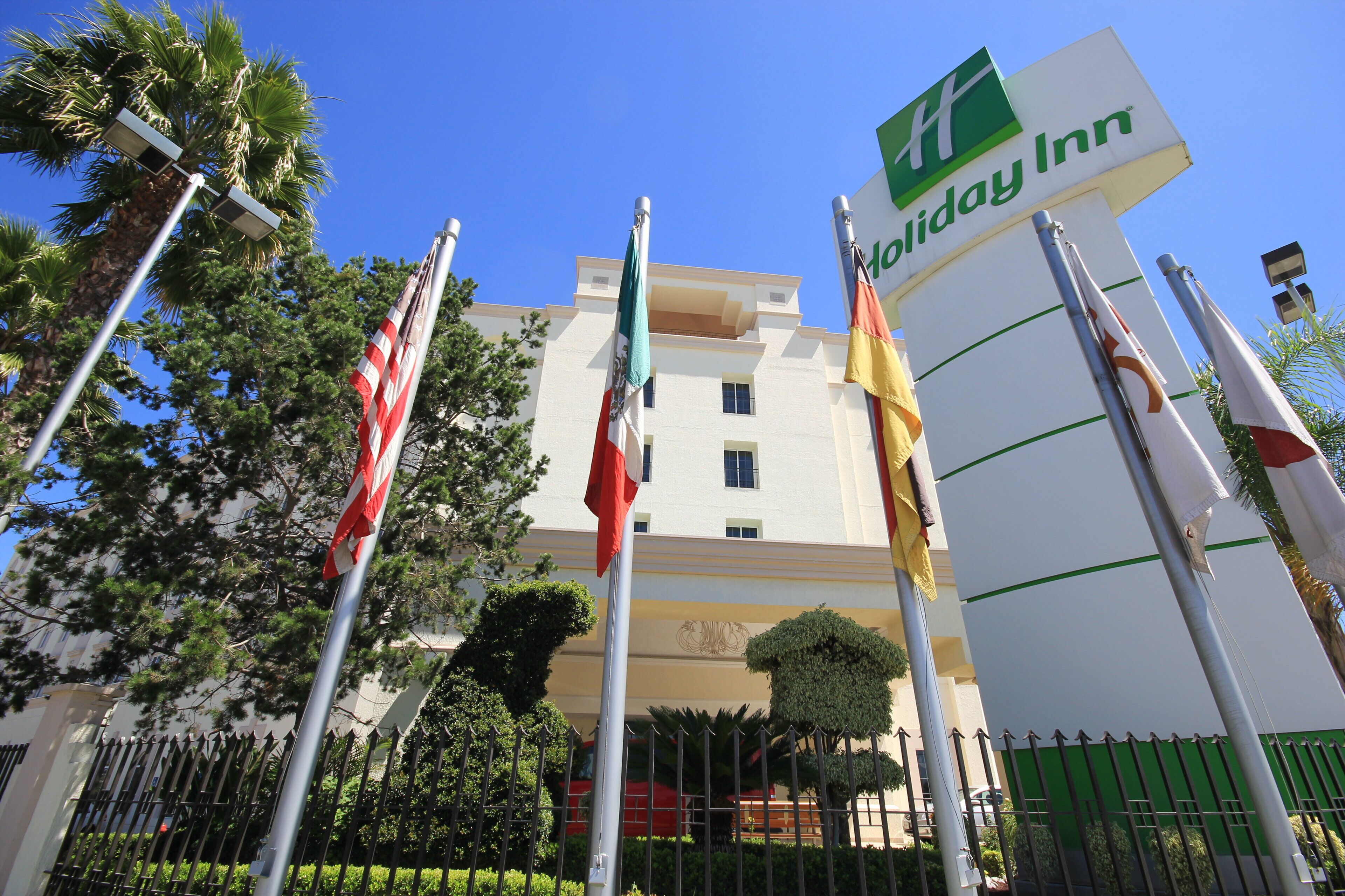 Un hotel Holiday Inn con múltiples banderas, un cielo azul despejado y un jardín bien cuidado.