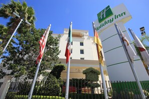 A Holiday Inn hotel with multiple flags, a clear blue sky, and a well-maintained garden.
