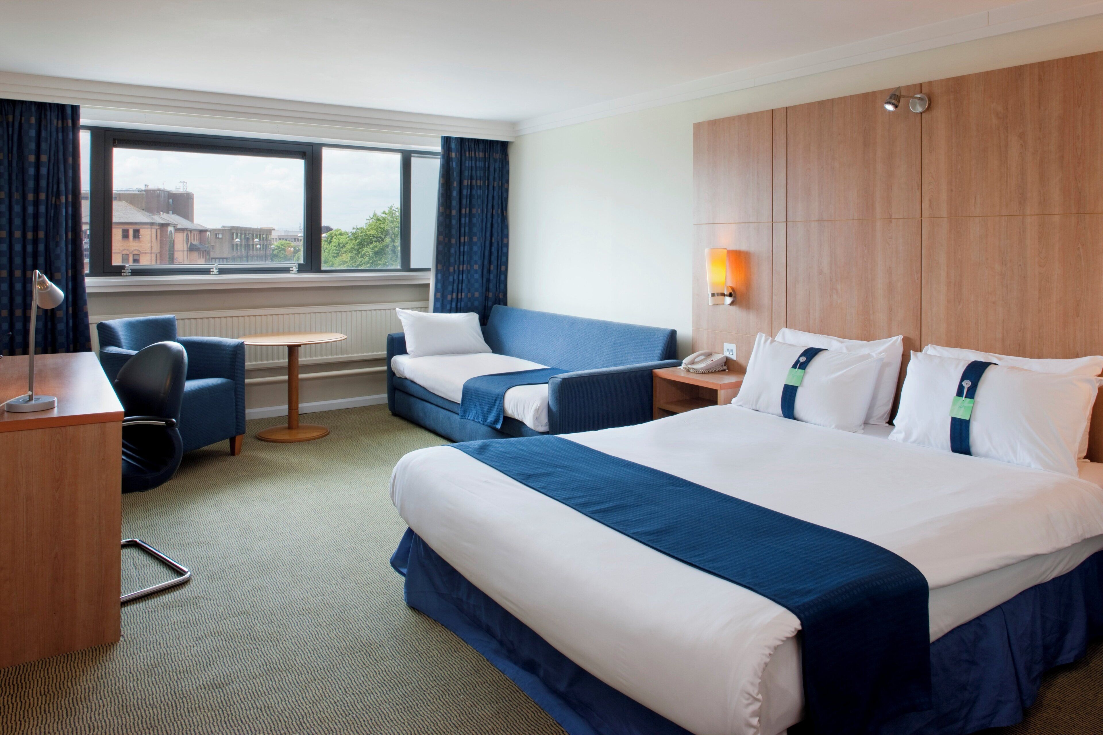 A hotel room with a large bed, two smaller beds, a desk, a chair, a table, a lamp, a phone, and a window with curtains.