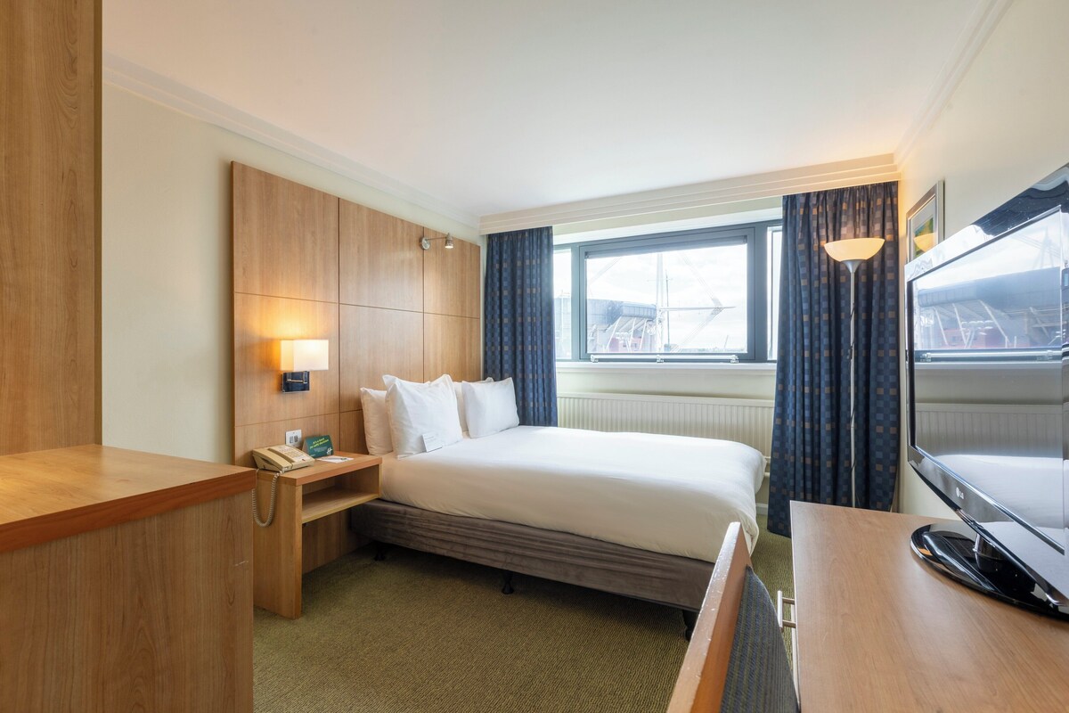 Une chambre d’hôtel avec un lit, un bureau, une lampe, une télévision et une fenêtre avec des rideaux.