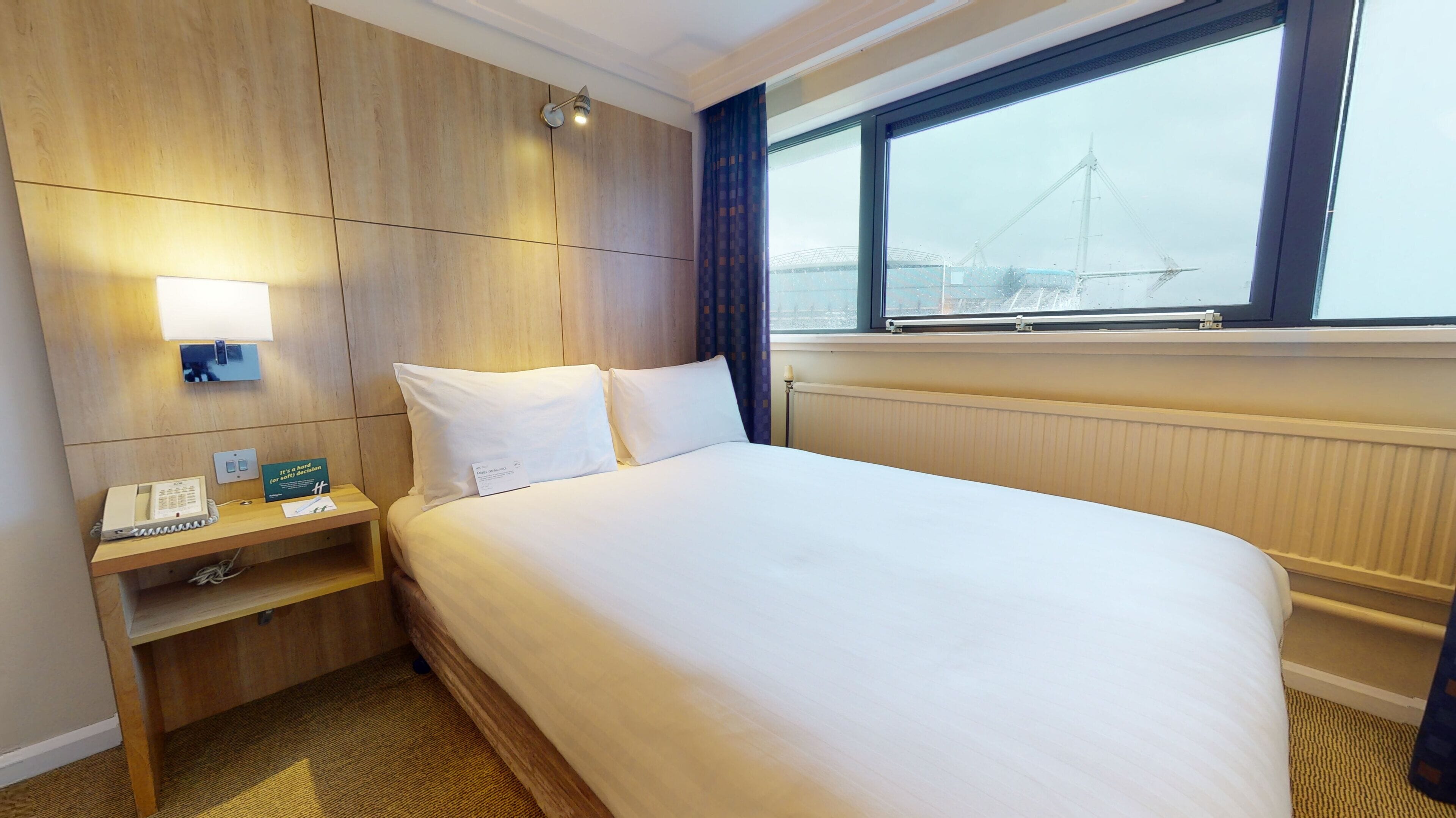 A hotel room with a bed, a nightstand, a telephone, a lamp, a window with a view of a bridge, and a radiator.