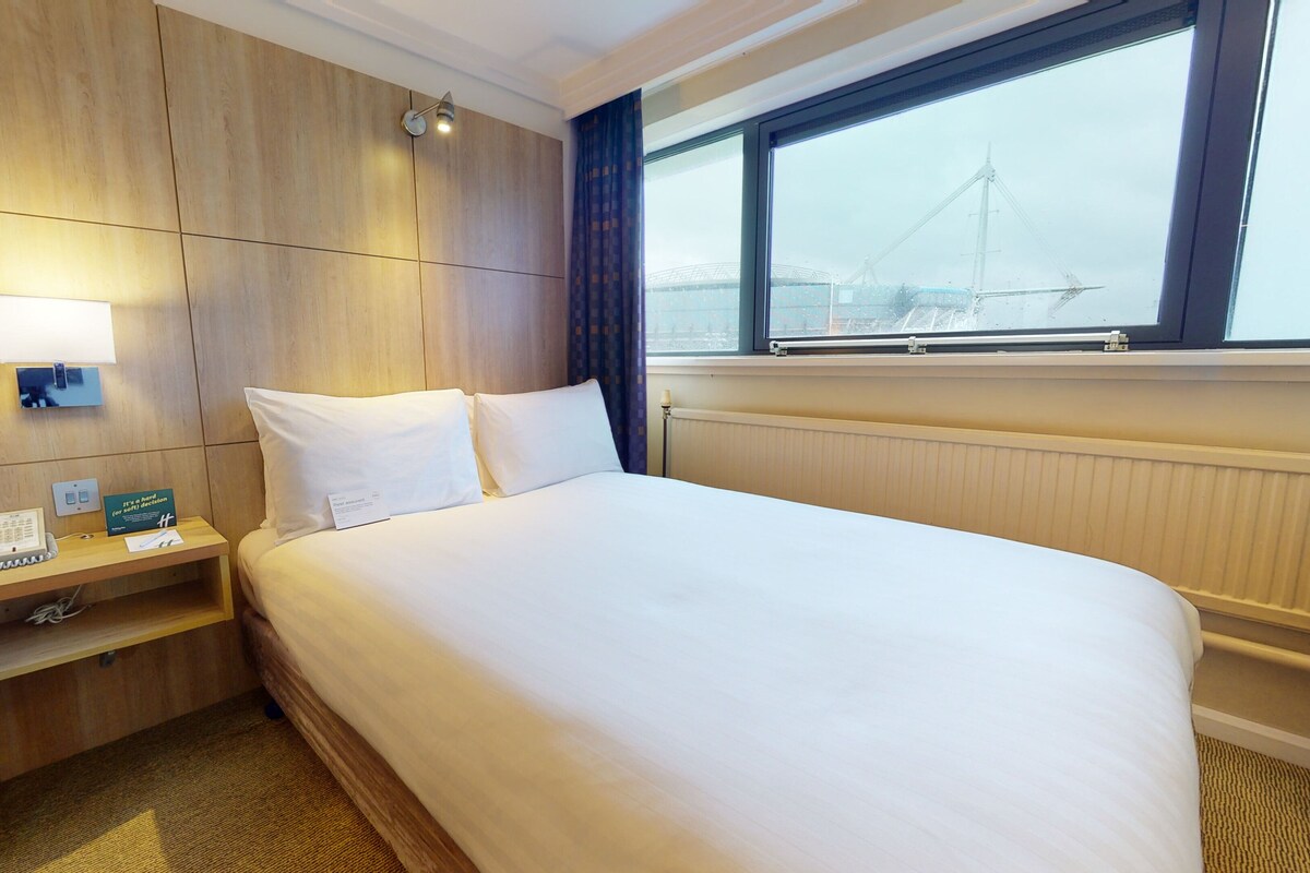 Une chambre d’hôtel avec un grand lit, une table de chevet avec un téléphone et un panneau, une fenêtre donnant sur un pont et un lampadaire fixé au mur.