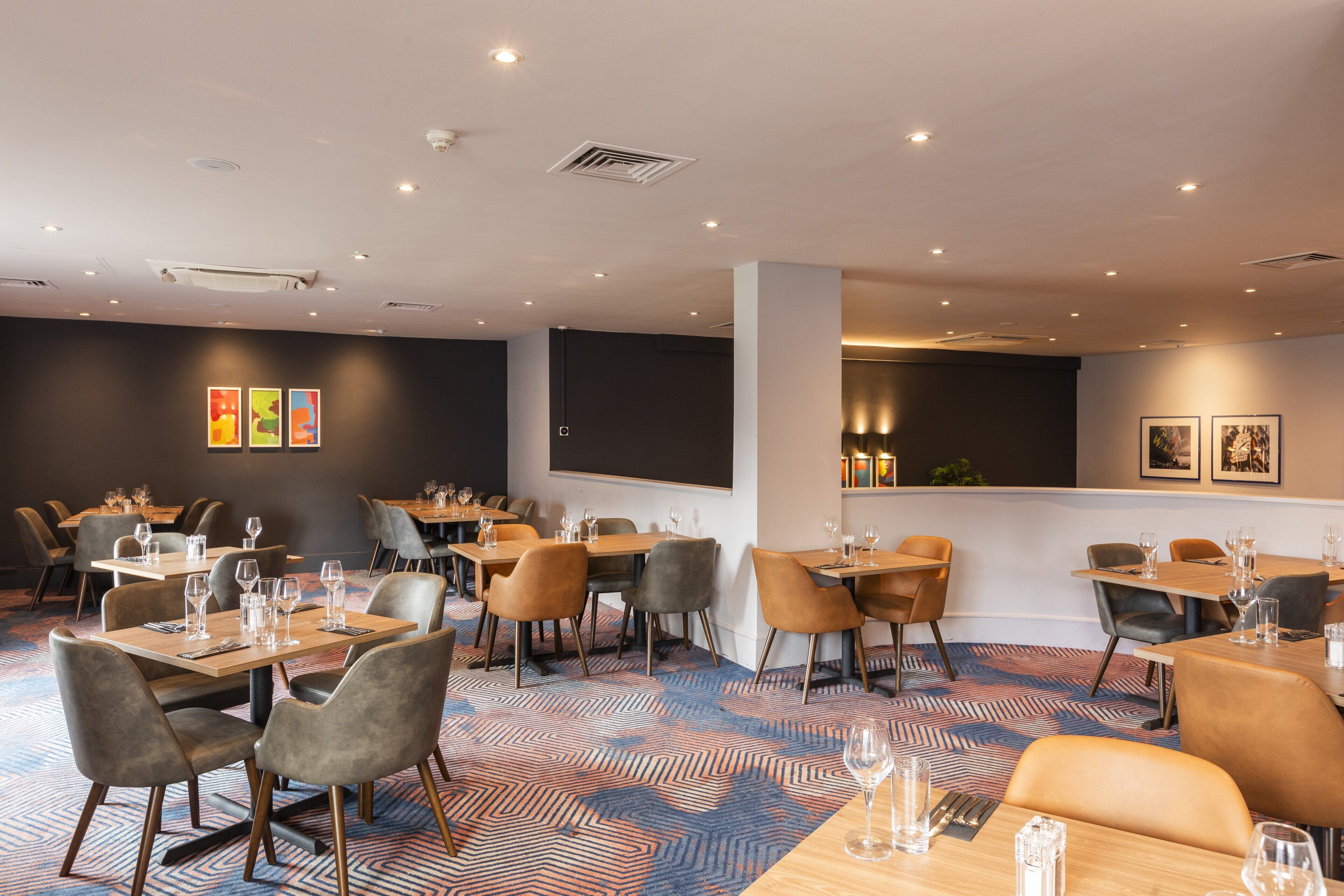 A modern restaurant with multiple tables and chairs, a patterned carpet, and artwork on the walls.