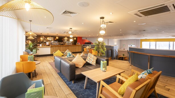 A modern hotel lobby with a bar area, seating arrangements, and a staircase leading to an upper level.