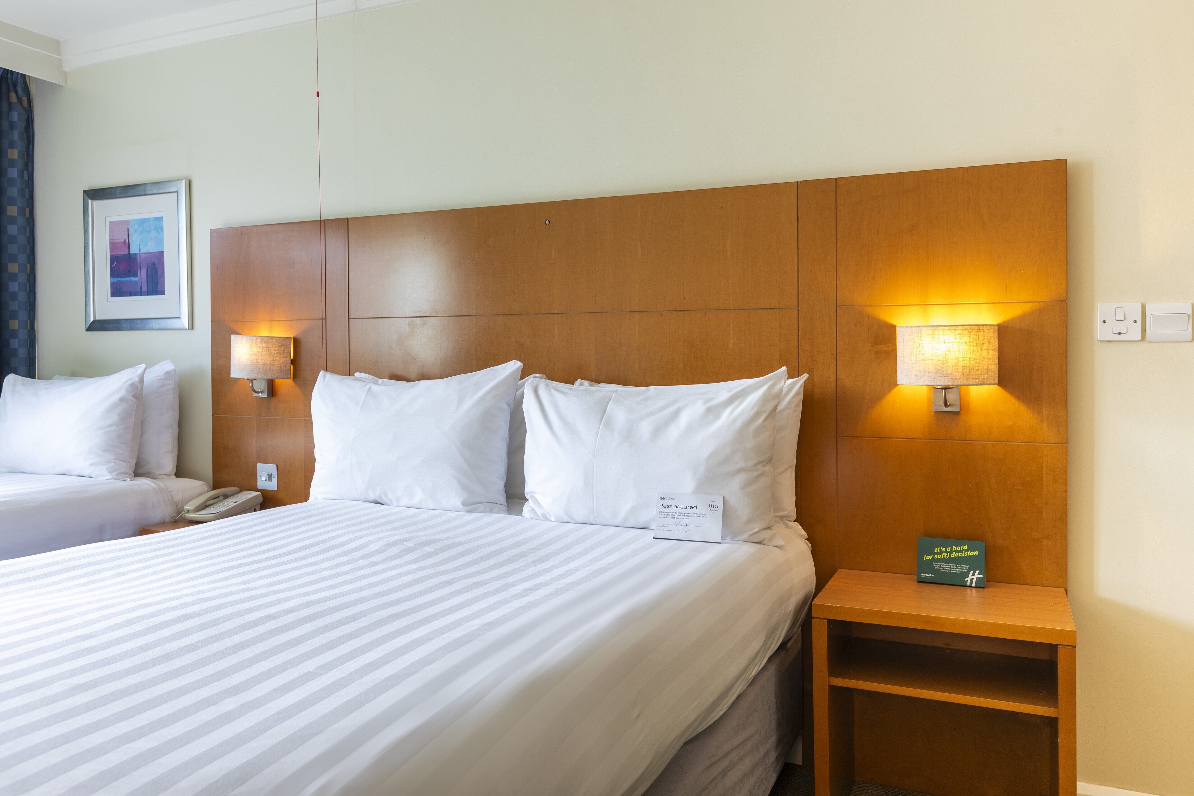 Ein Hotelzimmer mit zwei Betten, einem Holzkopfende, Nachttischlampen, einem Telefon, einem kleinen Tisch und einem gerahmten Bild an der Wand.
