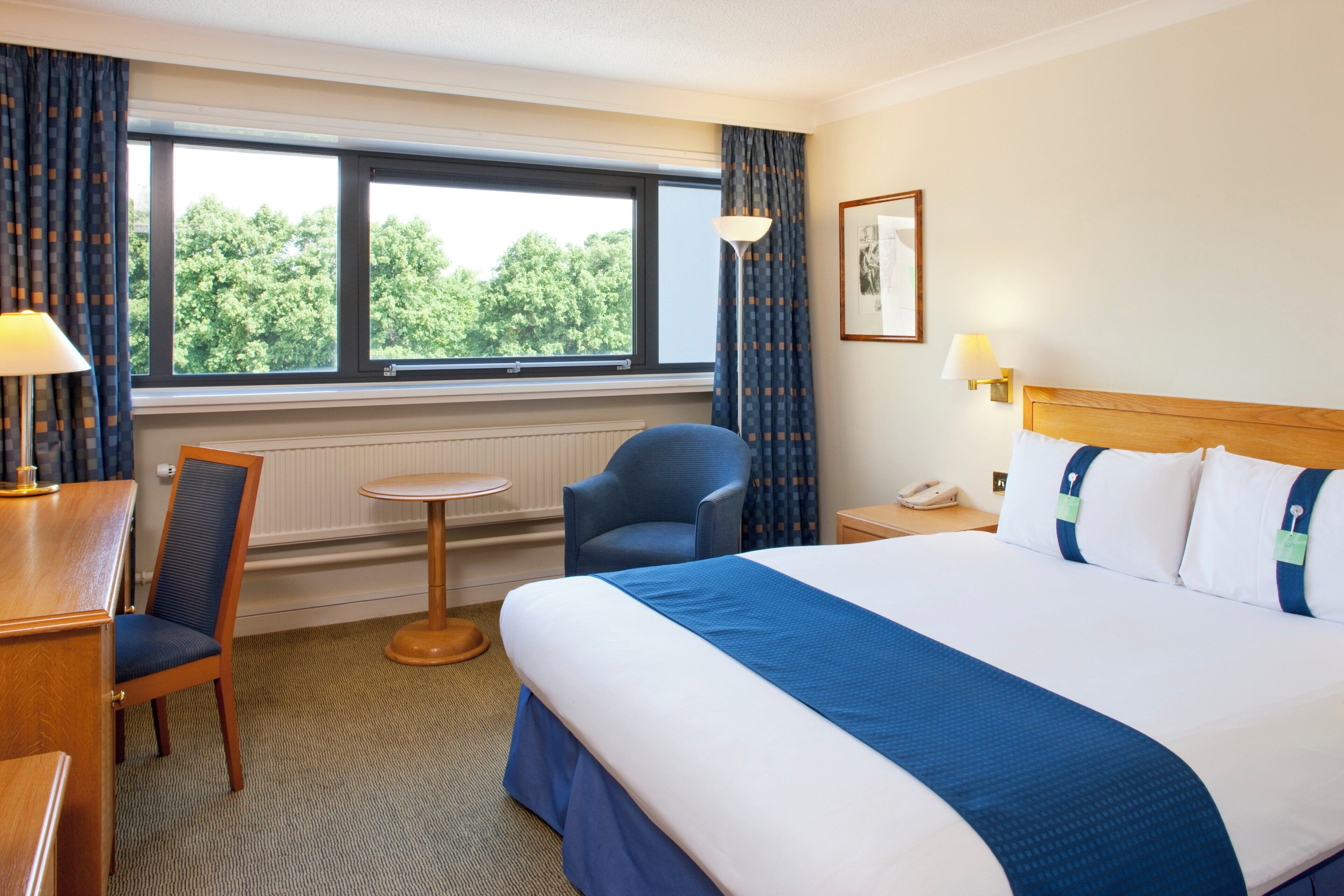 Une chambre d’hôtel avec un lit, un bureau, une chaise, une fenêtre avec des rideaux et une lampe.