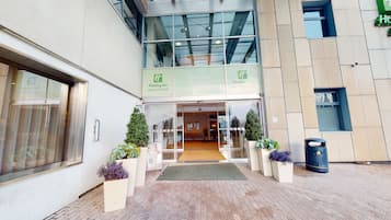 The entrance to Holiday Inn Cardiff City Centre with a clear glass facade, a welcoming sign, and potted plants at the entrance.