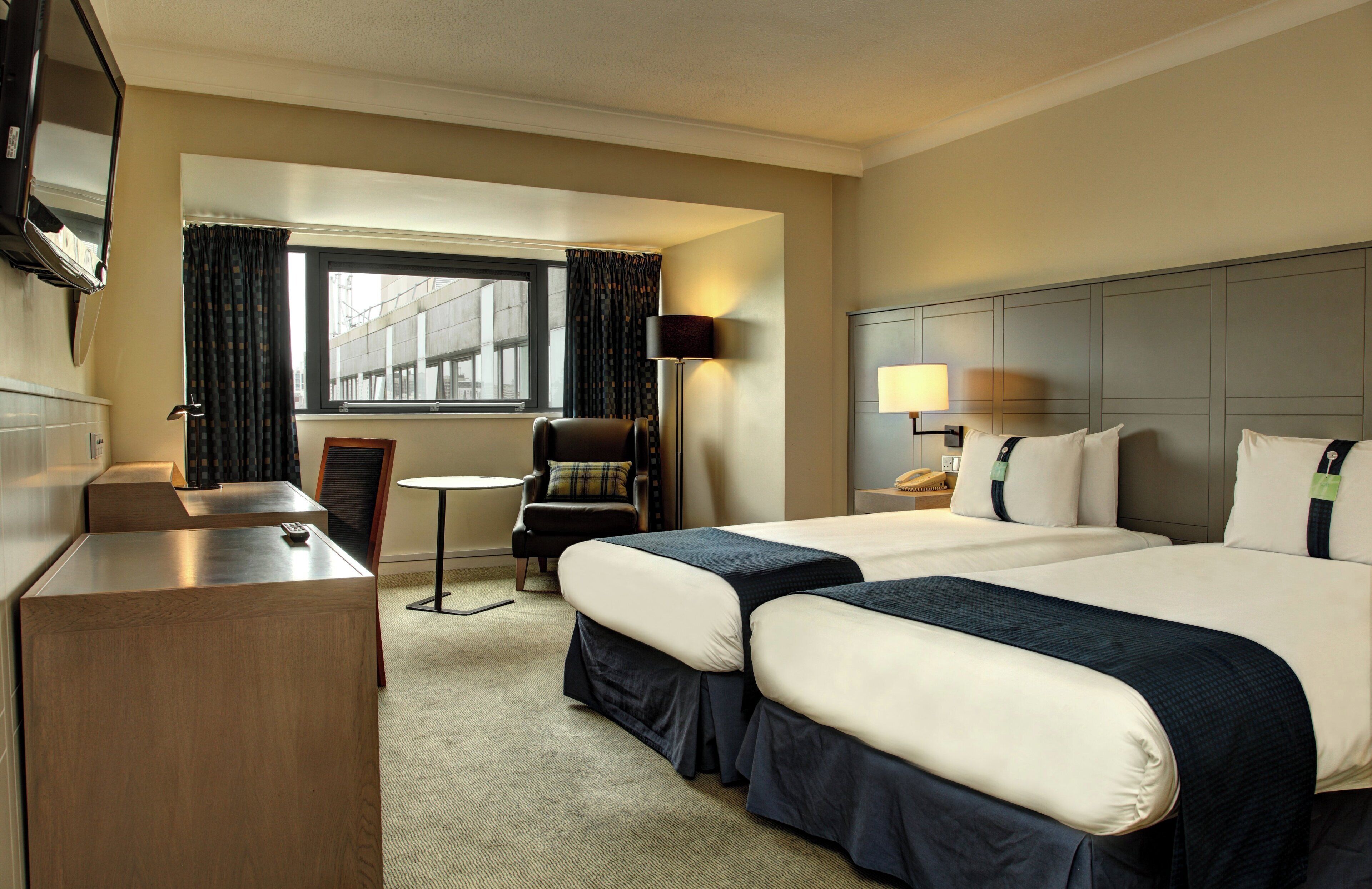 A hotel room with two beds, a desk, a chair, a small table, and a window with curtains.