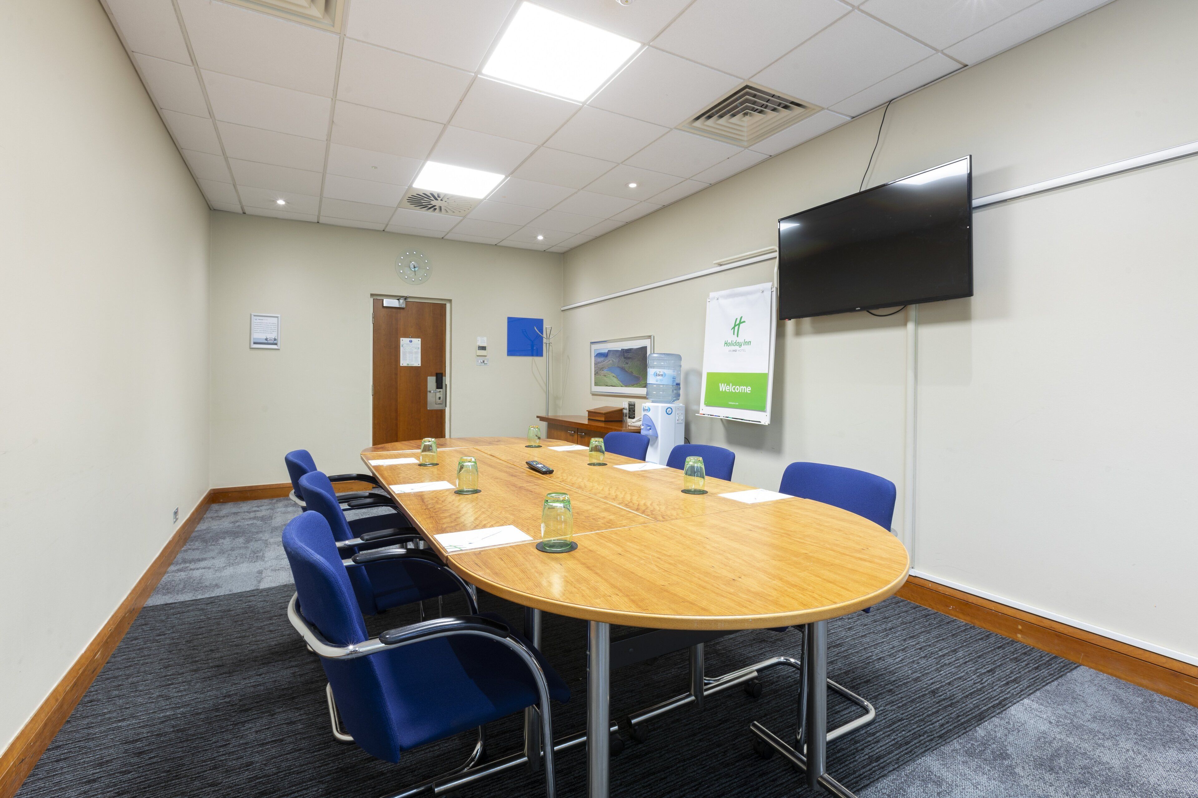 A conference room with a long table, blue chairs, a flat-screen TV, and a welcome sign.