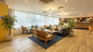 A modern hotel lobby with a wooden floor, a seating area with a table and chairs, and a bar in the background.