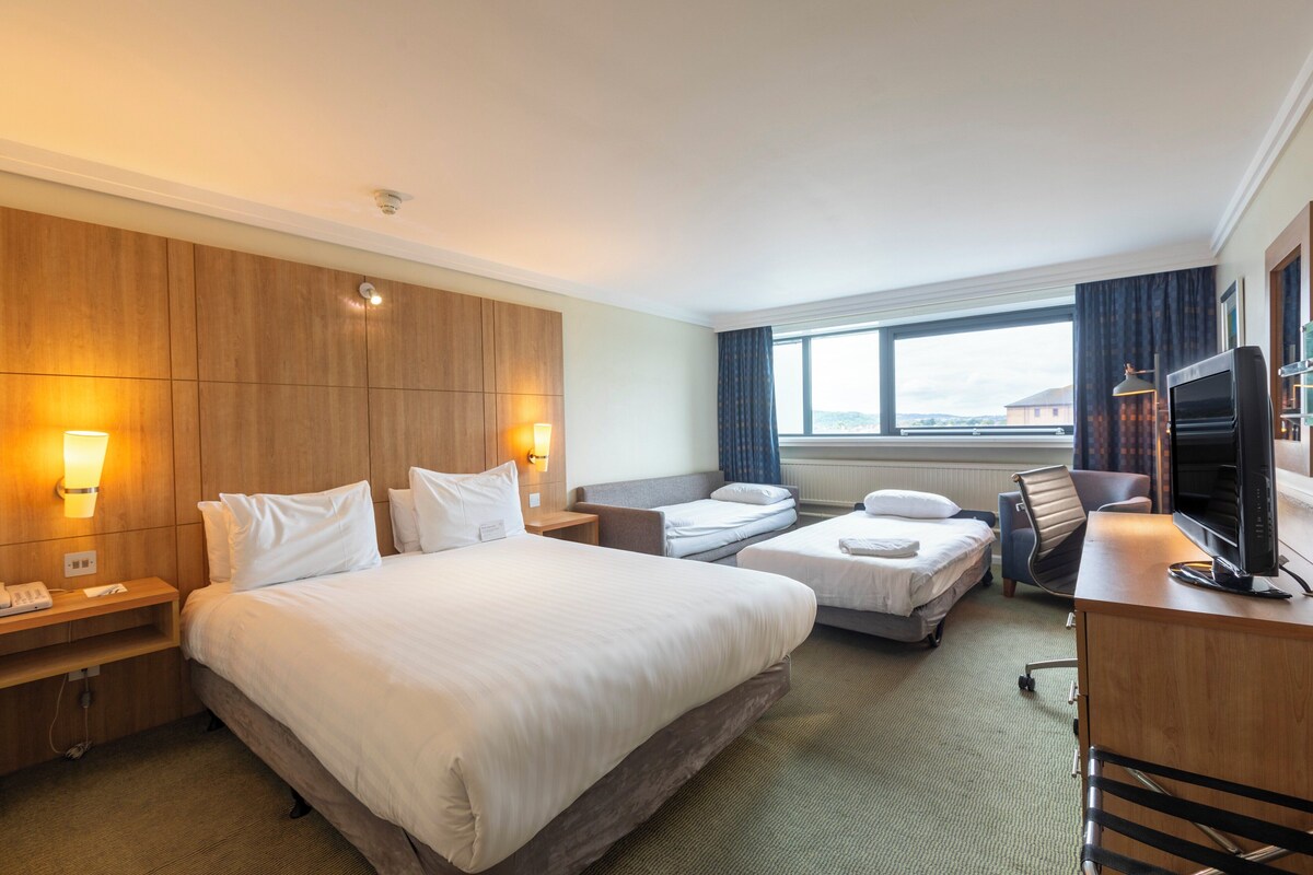 Une chambre d’hôtel avec un grand lit, un lit plus petit, un bureau avec une télévision et une fenêtre donnant sur un beau paysage.