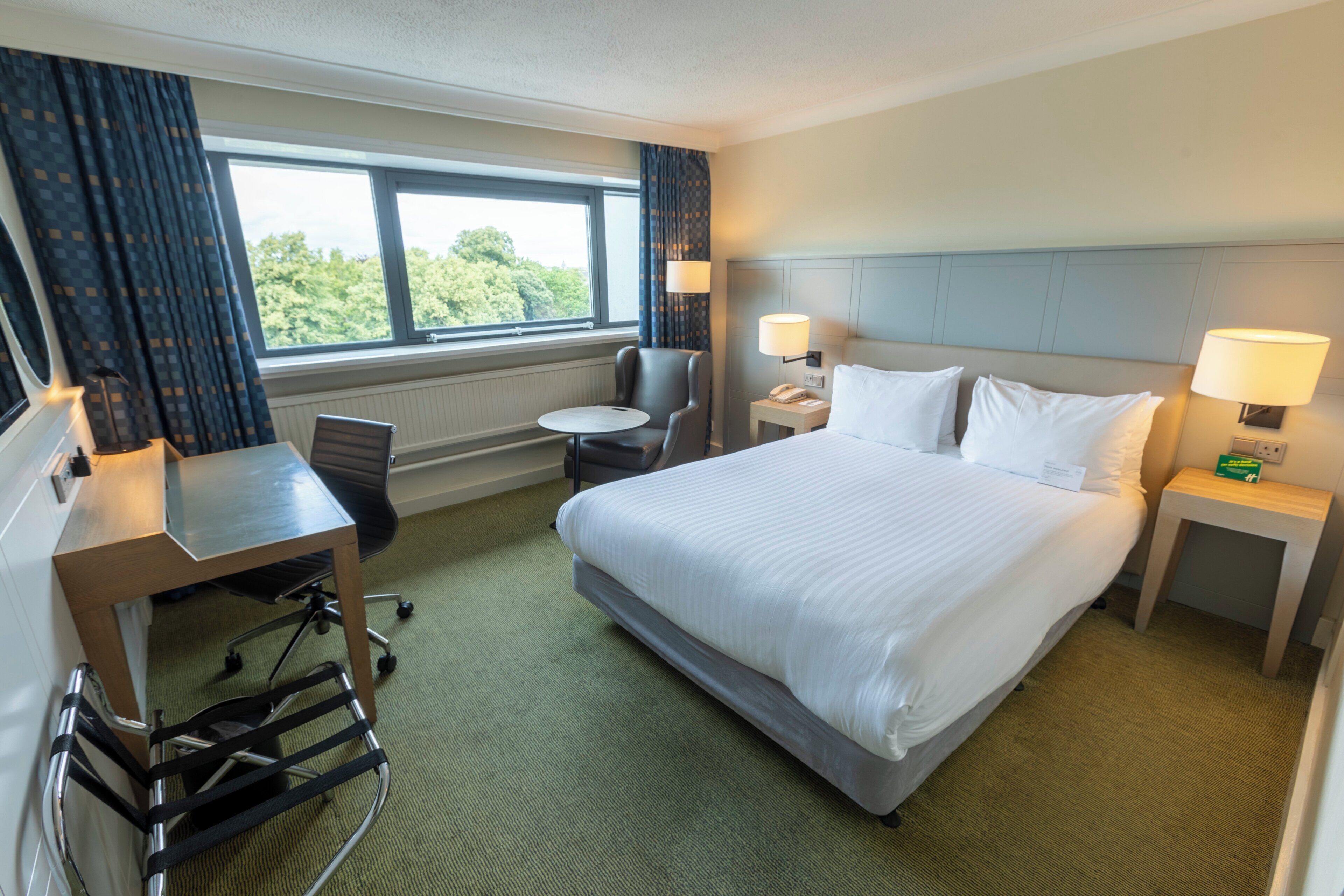 Ein Hotelzimmer mit einem großen Bett, einem Schreibtisch, einem Sessel, einem kleinen Tisch und Blick ins Freie durch das Fenster.