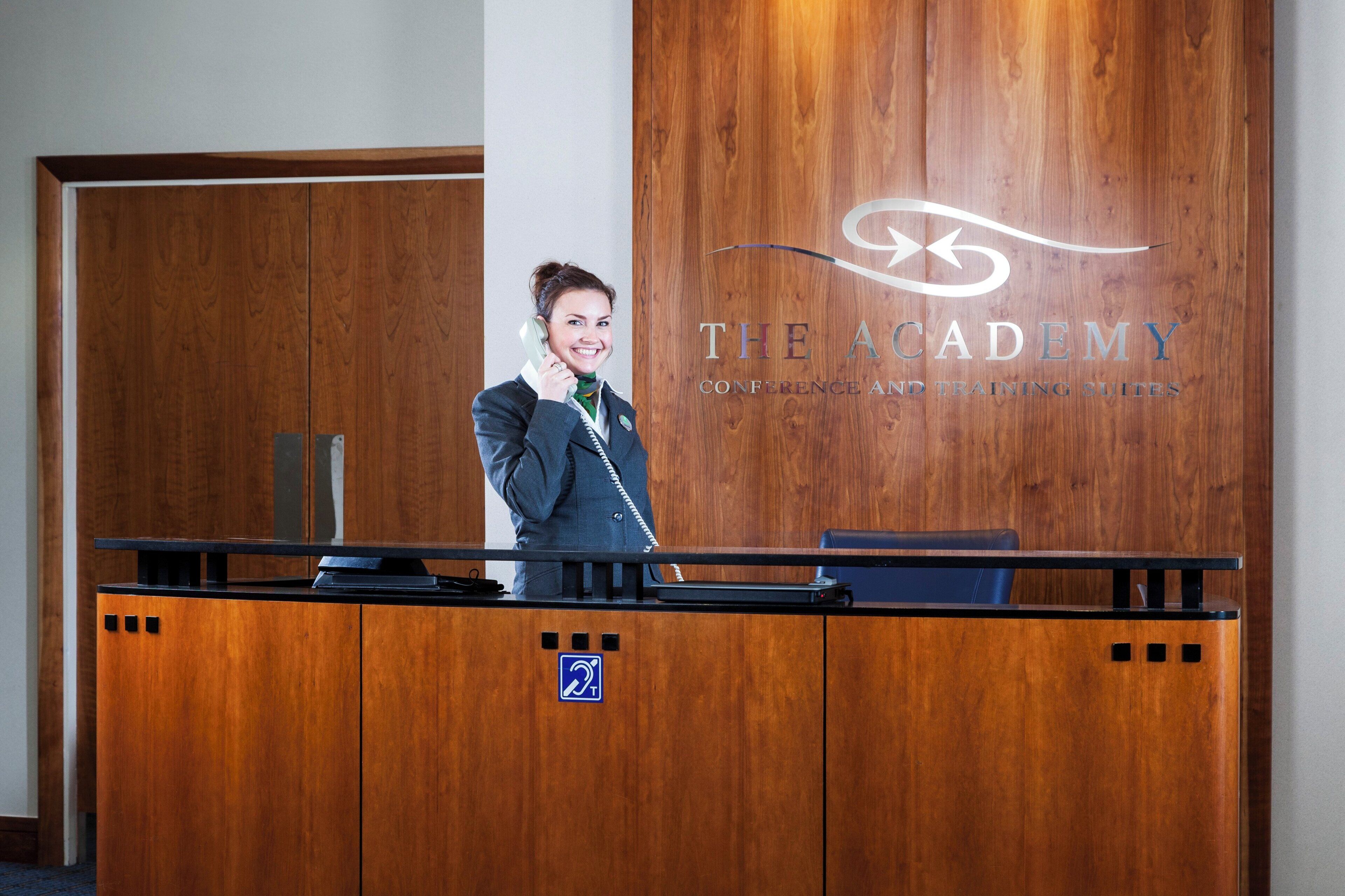 Een receptiebalie met houten afwerking, een logo voor 'The Academy Conference and Training Suites', en een blauw toegankelijkheidsbord.