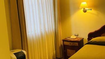 A hotel room with a bed, a bedside table with a lamp, a heater, and a window with curtains.
