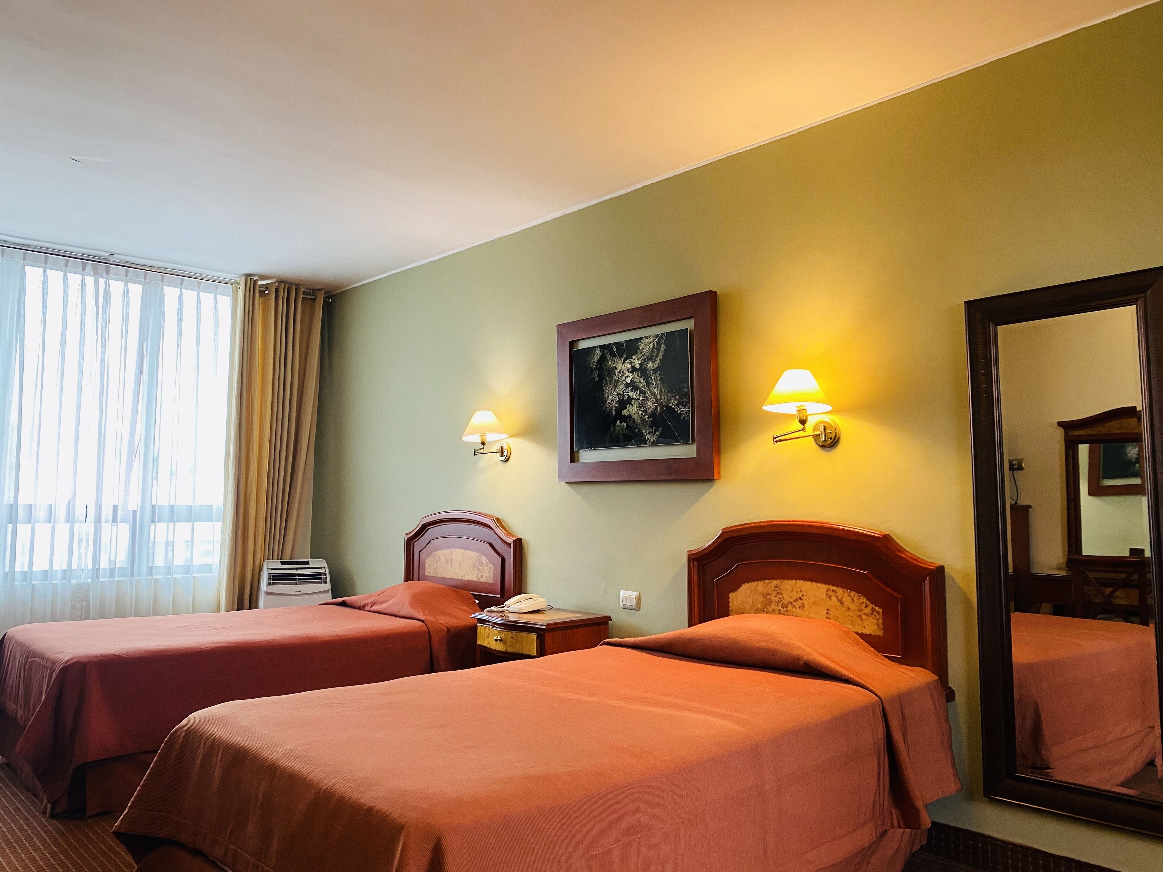 A hotel room with two beds, a bedside table with a lamp, a framed picture on the wall, and a large mirror.