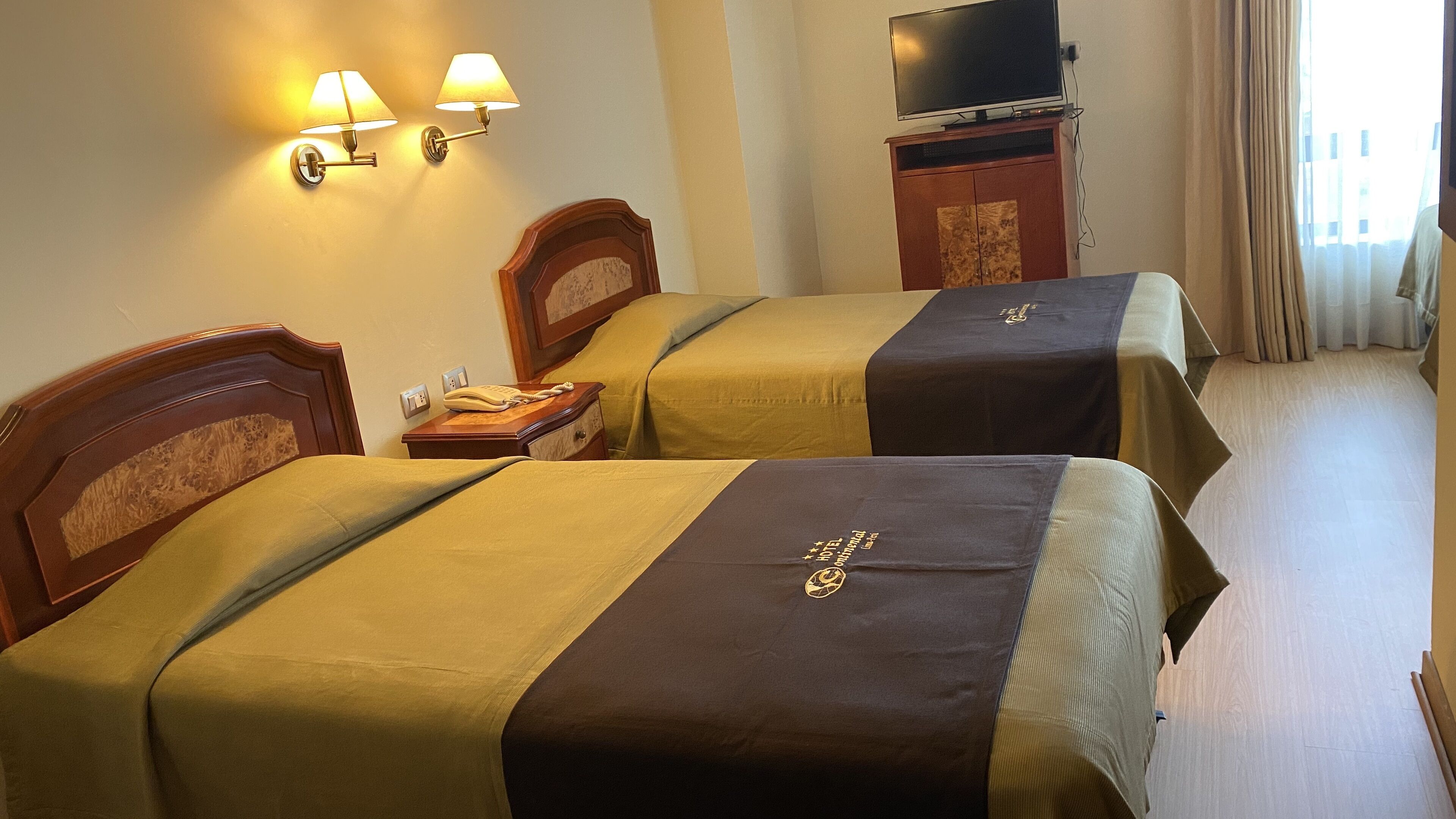 Dos camas individuales con cabeceras, un teléfono junto a la cama, un televisor sobre un soporte y una cortina en la ventana.