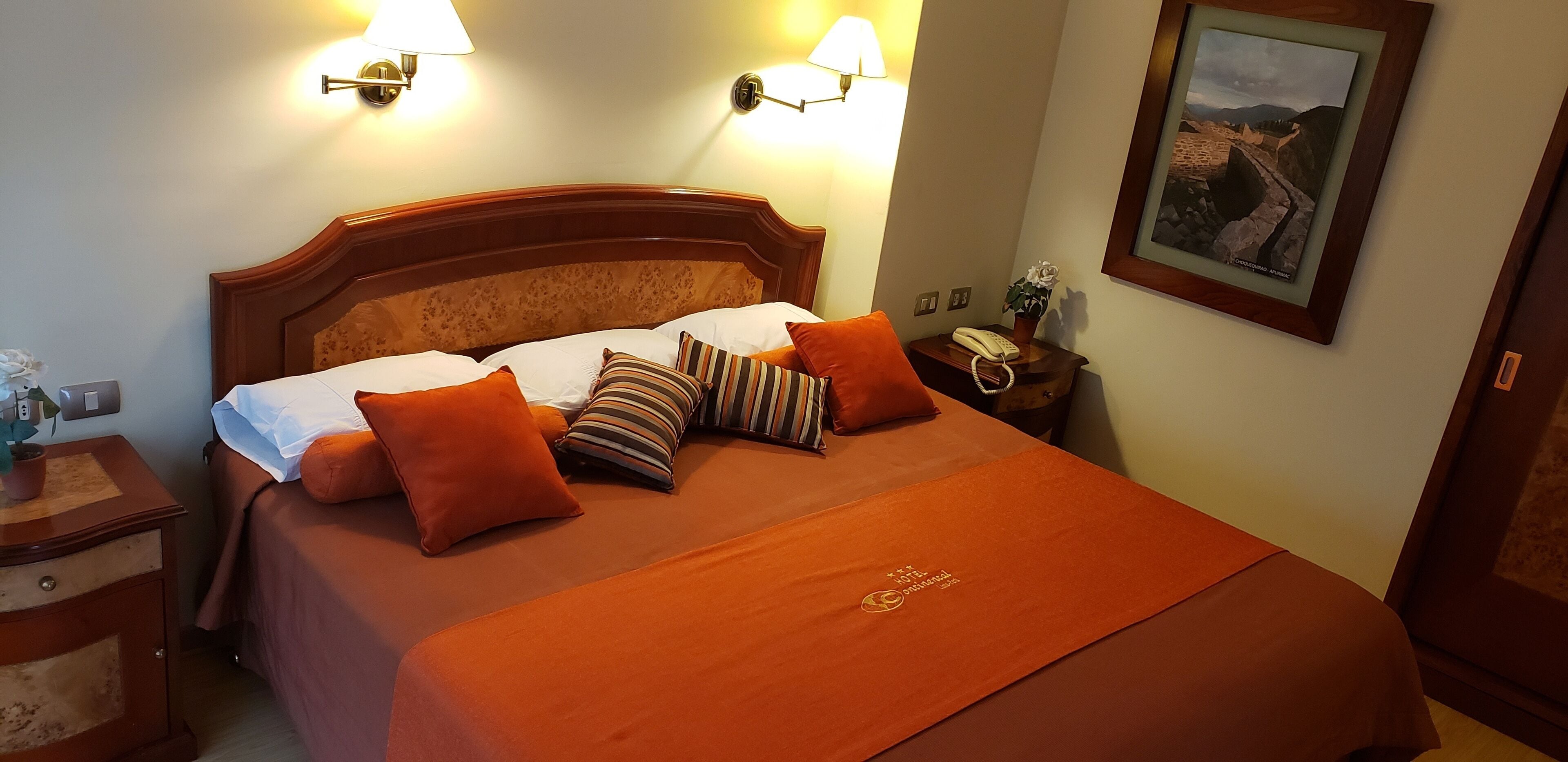Hotelkamer met houten hoofdbord, een bed met oranje beddengoed, twee bedlampen, een nachtkastje met een telefoon en een ingelijst foto aan de muur.