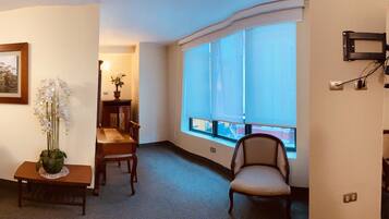 A hotel room with a bed, a desk with a chair, a potted plant, a window with a view, and a wall-mounted TV.