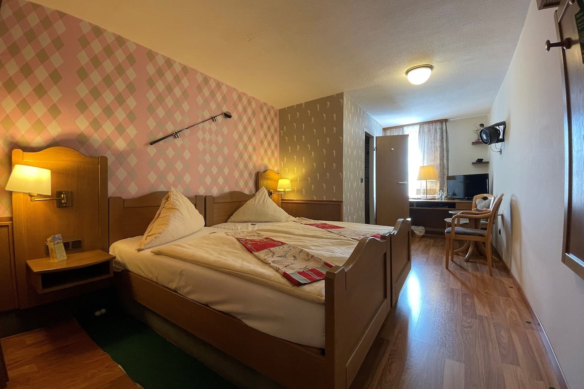 A hotel room with a bed, bedside lamps, a desk with a chair, a television, and a patterned wall.