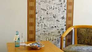 A wooden table with a bottle and a bowl of snacks, a chair with a patterned cushion, and a wall with a newspaper print design. - Bebras Hessischer Hof (Bebra)
