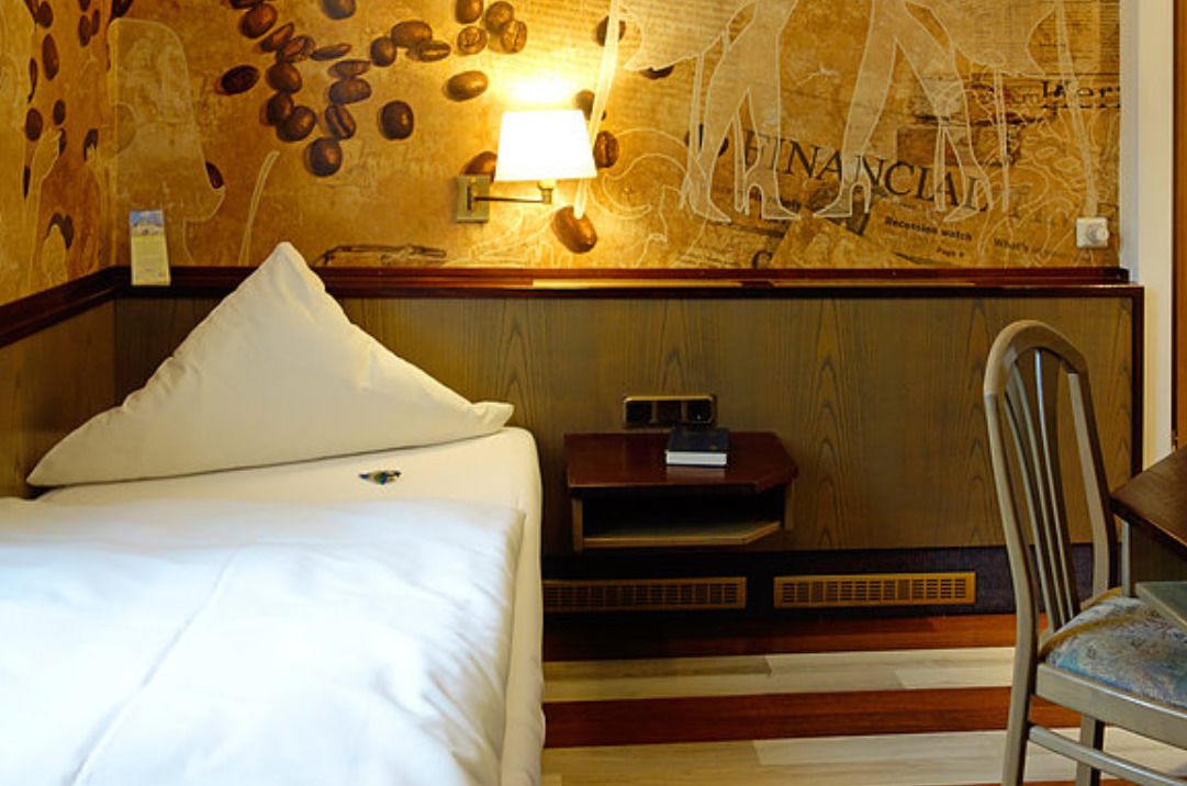 A hotel room with a bed, a nightstand with a lamp, a chair, and a wall with a map design.