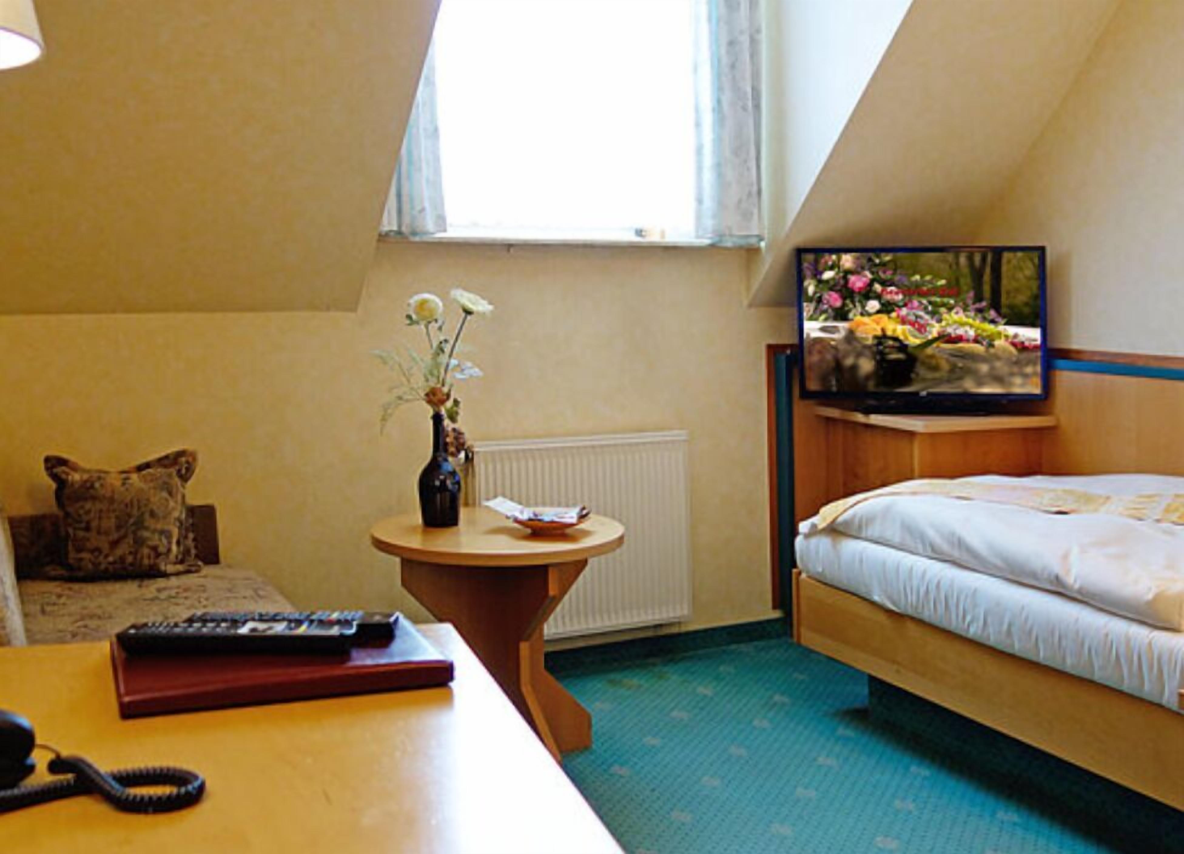 A hotel room with a bed, a desk with a telephone and remote control, a small table with a vase of flowers, and a television on the wall.