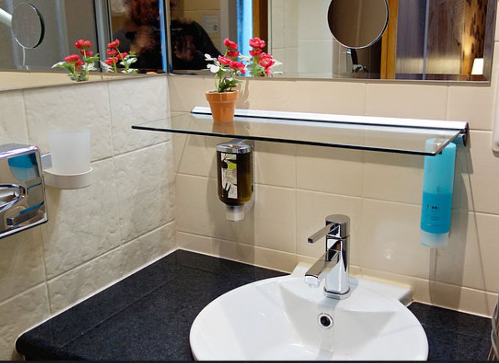 Ein Badezimmer mit einem weißen Waschbecken, einem Glastischchen, einem Spiegel und einer Topfpflanze mit roten Blüten.