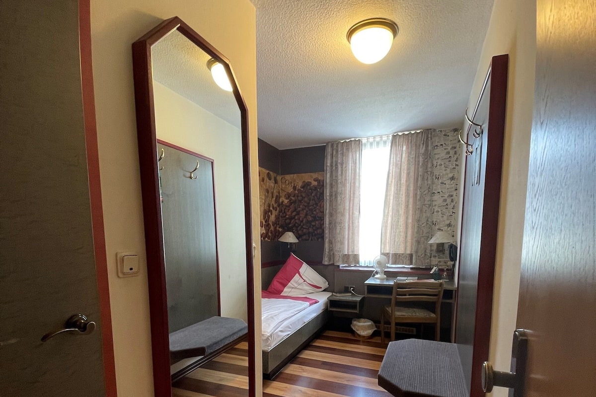 A compact hotel room with a wooden floor, a bed with a red and white blanket, a small desk with a chair, a mirror, and a ceiling light.