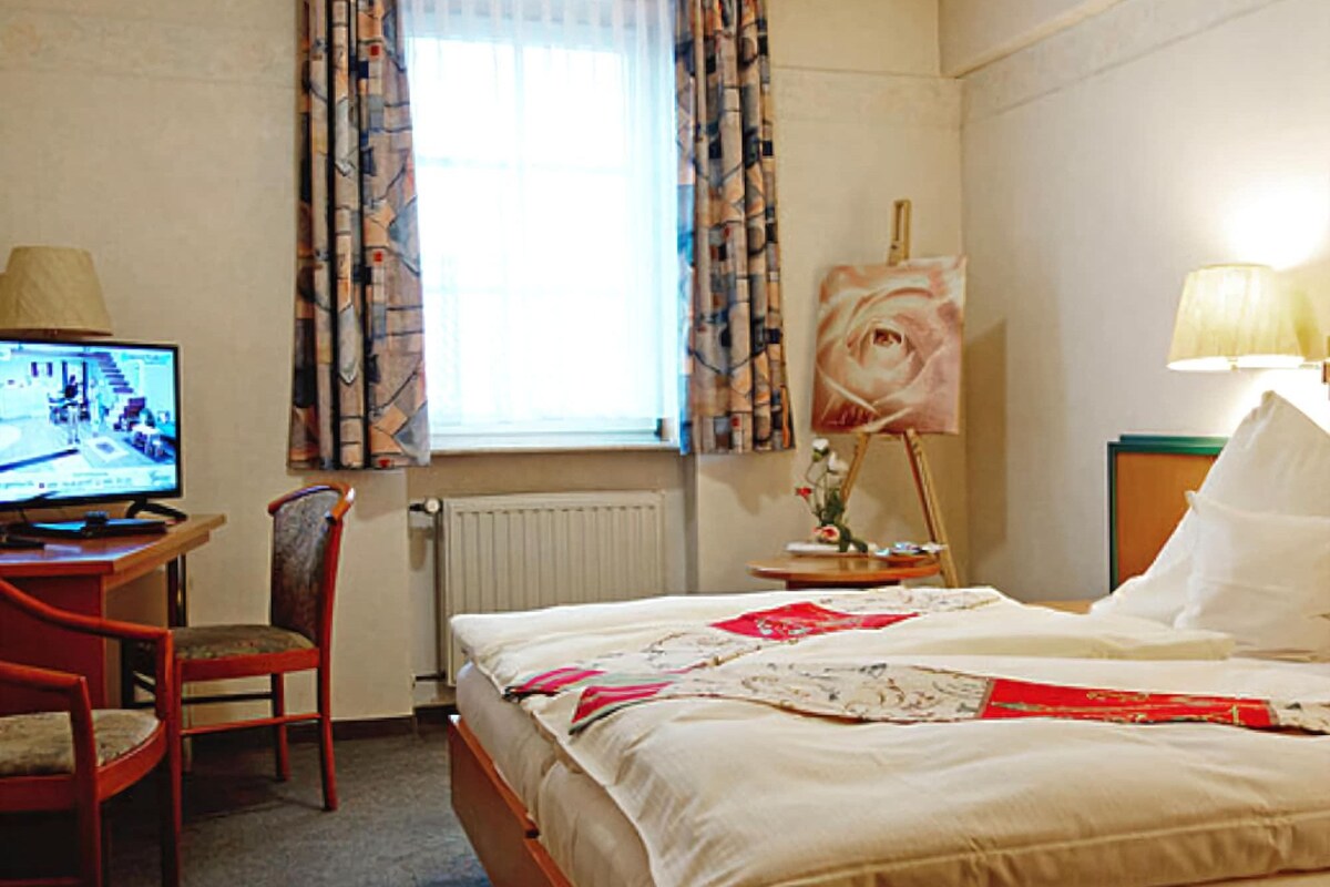 Habitación de hotel con cama, escritorio con computadora, silla, ventana con cortinas y un cuadro en la pared.